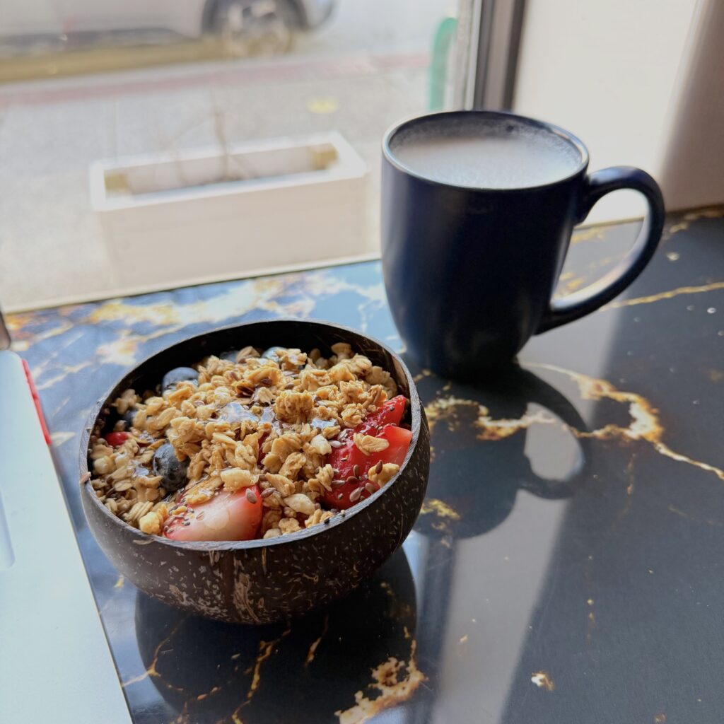 Mug of coffee and traditional acai bowl with granola, blueberries, and strawberries placed on top of a bar table in front of a large glass cafe window.
