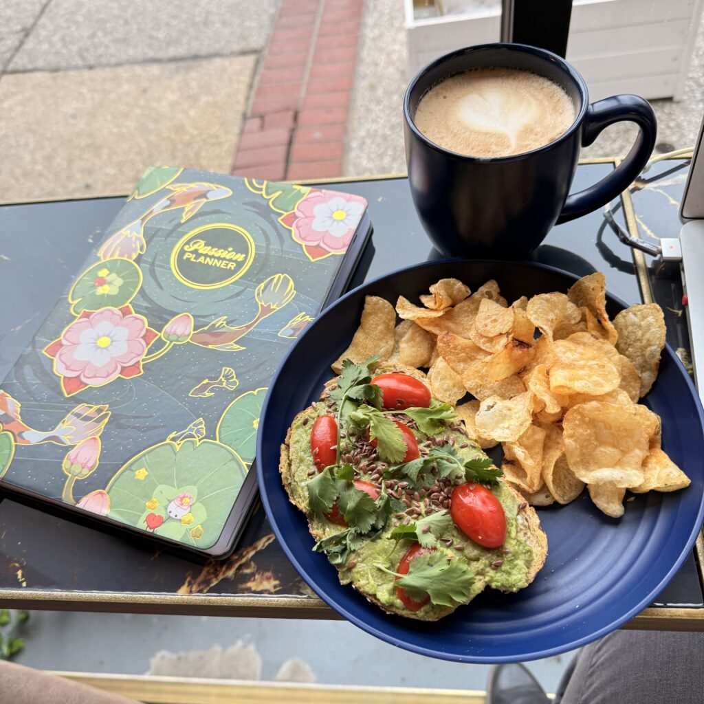 Bar table in front of a window with a mug of coffee, a planner, and plate of avocado toast and chips on it