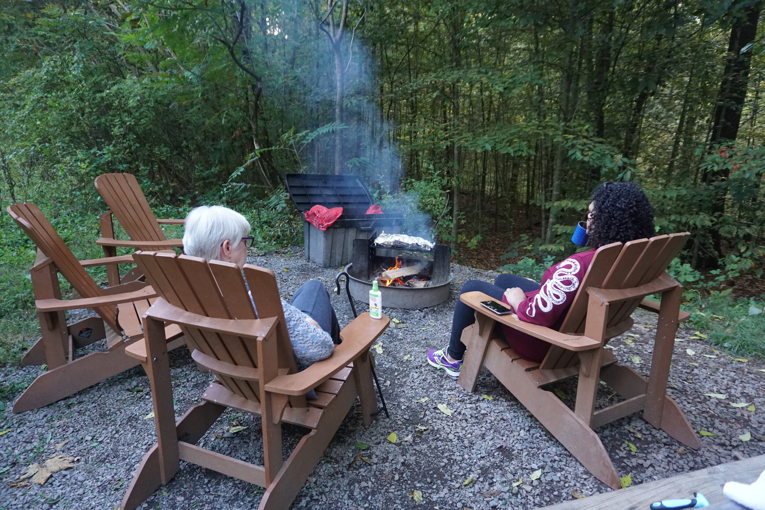 Two women sitting outside in lounge chairs surrounding a grill in the woods