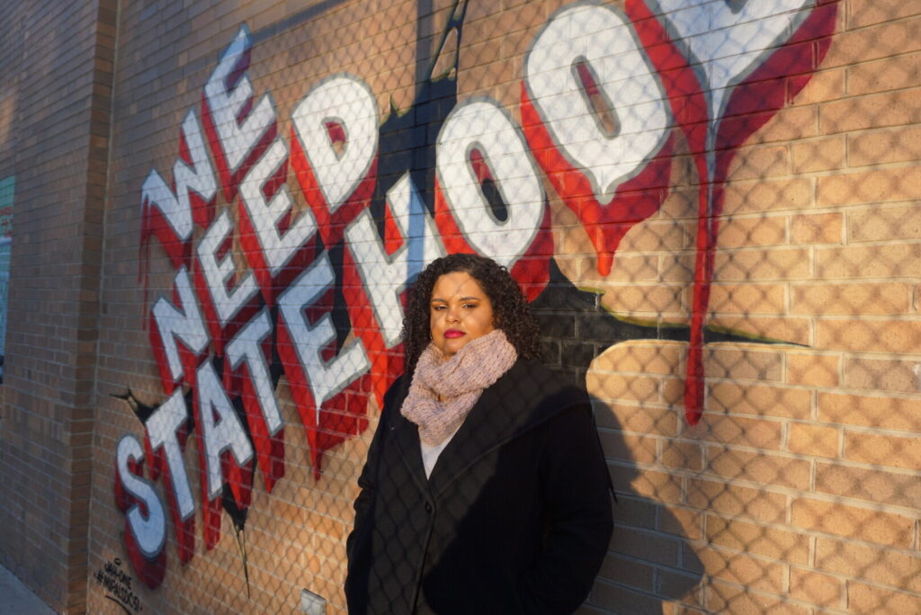 Woman standing in front of brick wall mural that says We Need Statehood