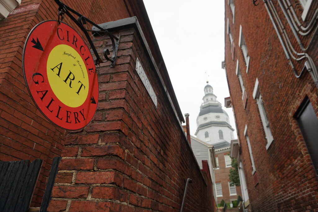 An art gallery sign on a brick building with a historic state capitol house in the background in Annapolis Maryland