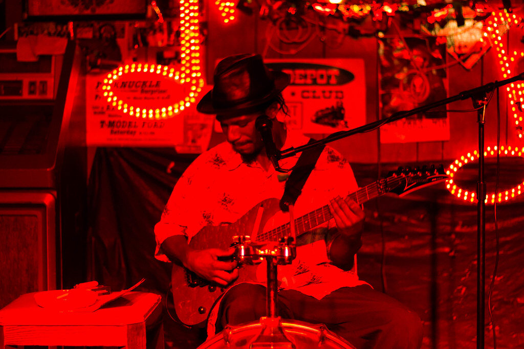 A blues musician playing guitar in a blues club lit up by fluorescent red lights.