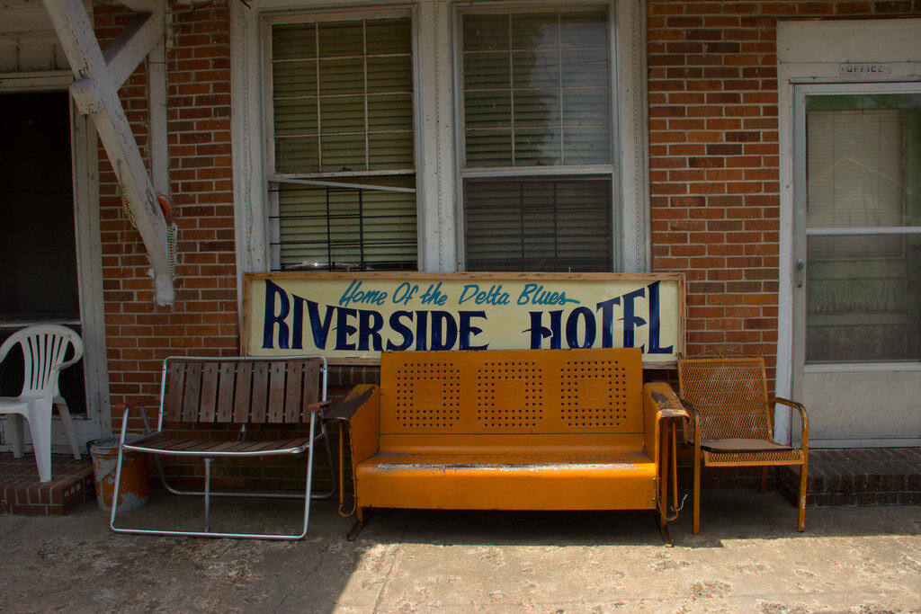 The porch of a brick house with a vintage sign reading Riverside Hotel and various dirty chairs.