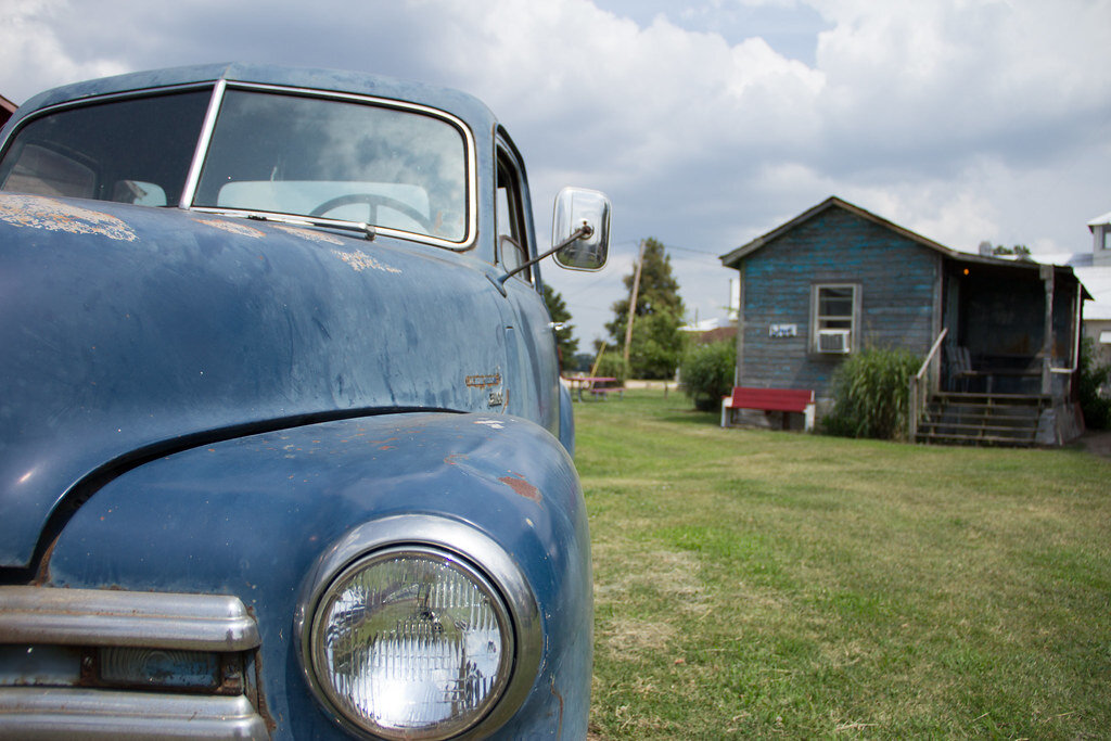 Antique blue buggie car on a grassy field with a blue wooden shotgun house in the background