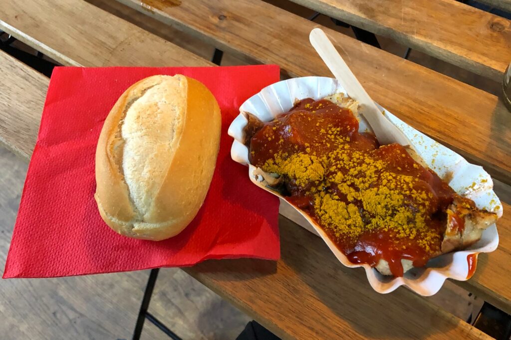 A bread roll sitting on a red napkin beside a paper bowl of cut up currywurst
