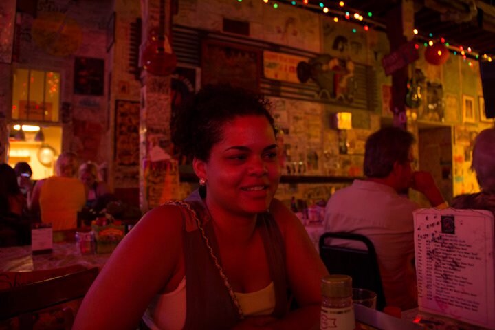 Smiling woman sitting in a blues bar with busy walls and multicolored string lights along the ceiling.