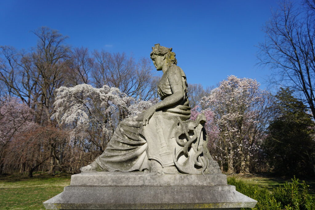 Statue of a sitting woman on a pedestal in a green lawn with magnolia trees in the background