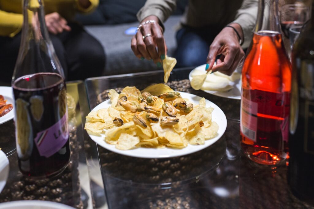 Plate of marinated mussels on top of potato chips with woman's hands picking some up on a table with two bottles of wine