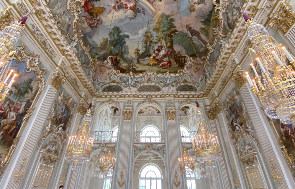 Elaborate ballroom with white walls and ornate gold accents, chandeliers, arched doorways, and a balcony in a Munich Germany palace