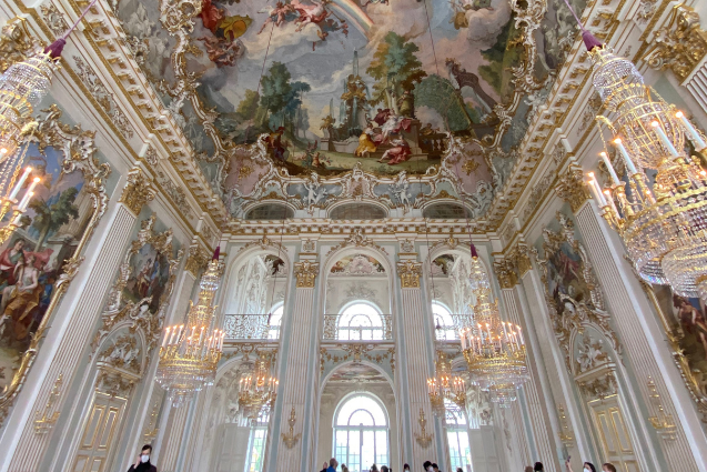 Elaborate ballroom with white walls and ornate gold accents, chandeliers, arched doorways, and a balcony in a Munich Germany palace
