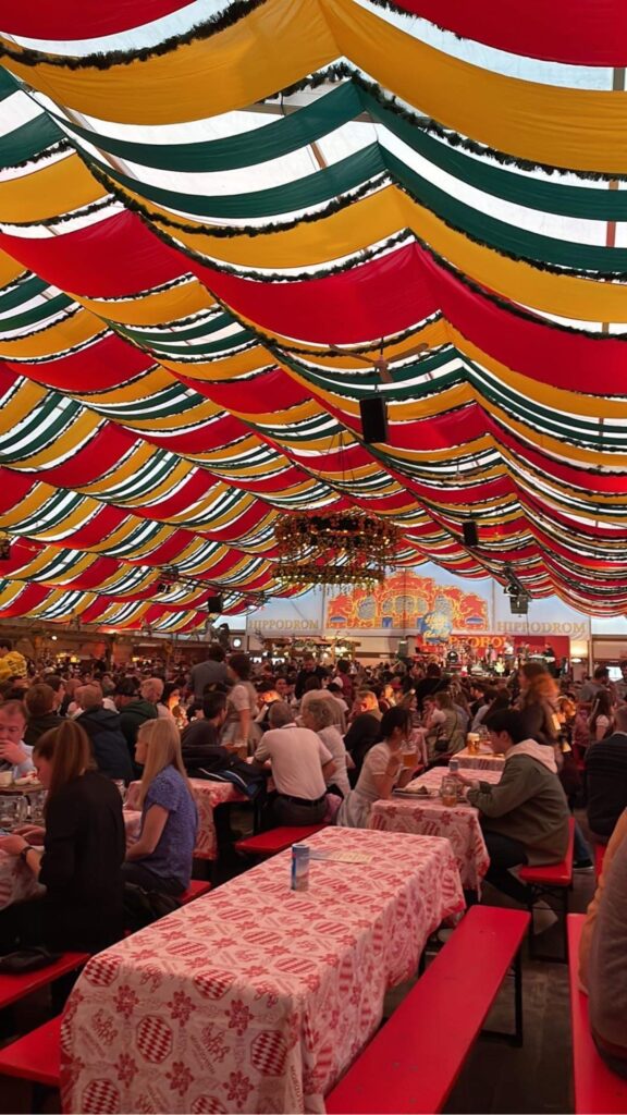 Inside of a beer tent with people sitting on long tables and benches, red, yellow, and green streamers above, and a medieval-style chandelier in the center.