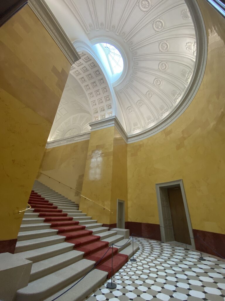 Palace staircase with red carpet, detailed white dome above, and yellow walls