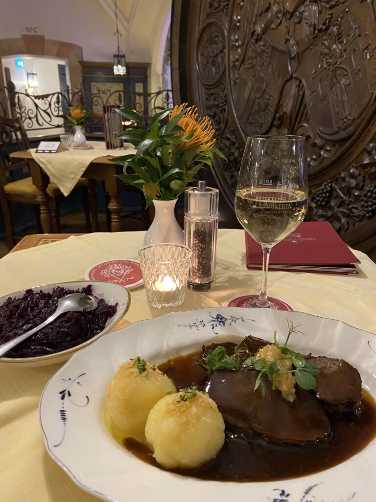 Plate of Bavarian food and a glass of white wine on a restaurant table with a large wooden carved barrel beyond it