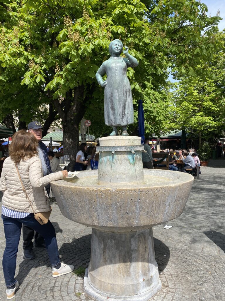 Statue of a woman in a fountain with people standing beside it drinking wine