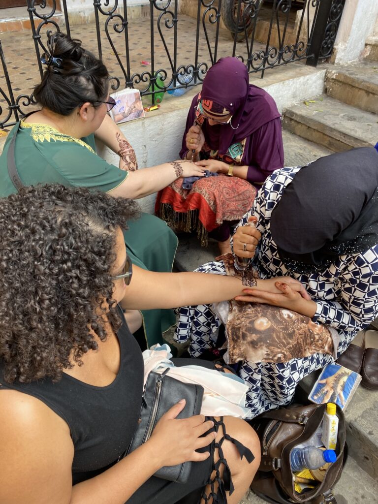 Two women sitting down getting henna done by two Moroccan women