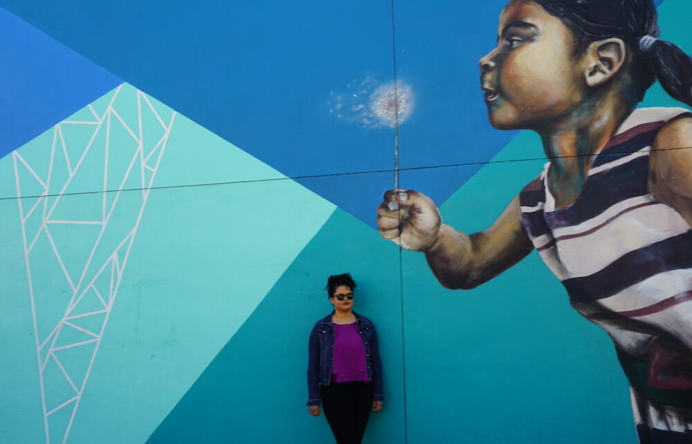 Woman standing in front of a large street mural of a little girl blowing a flower