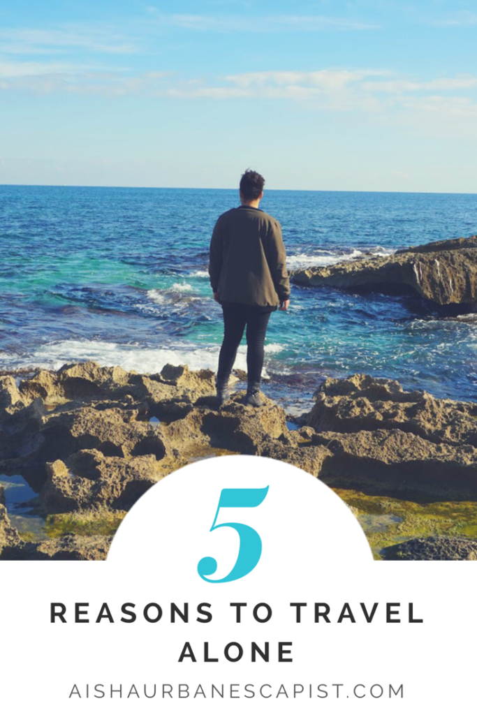 Image of woman standing on rocks in front of an ocean and the text five reasons to travel alone