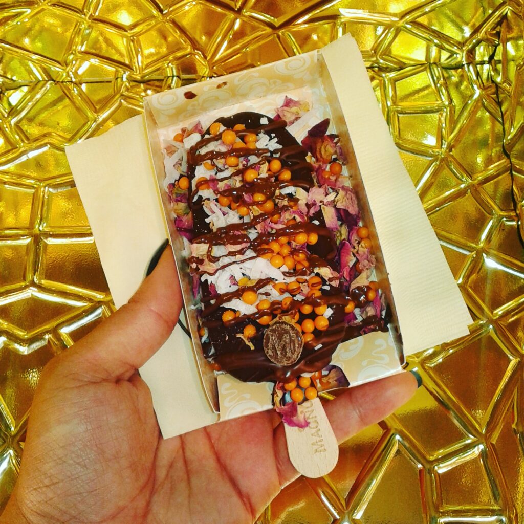 Hand holding a chocolate covered ice cream bar with colorful toppings against a gold background