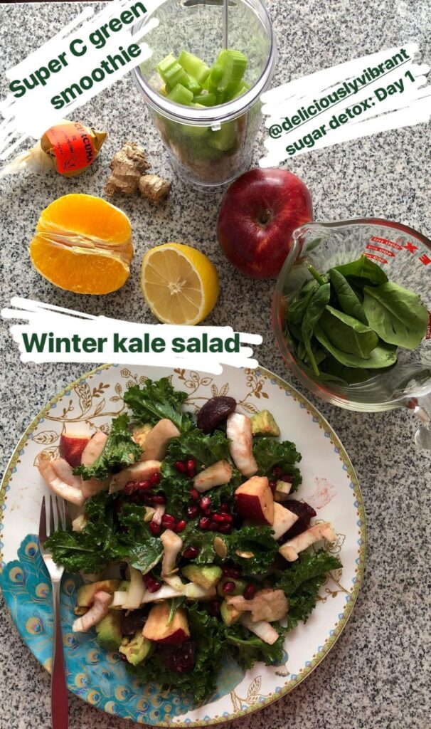 Flat lay showing a plate of winter kale salad, a measuring cup filled with spinach, a smoothie cup with unblended ingredients in it, and a cut lemon, orange, apple, and ginger root.
