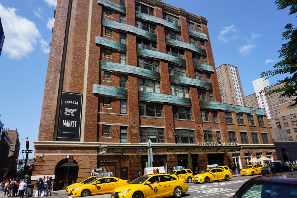 Brick building with green design elements on a city street with multiple yellow taxis driving by.