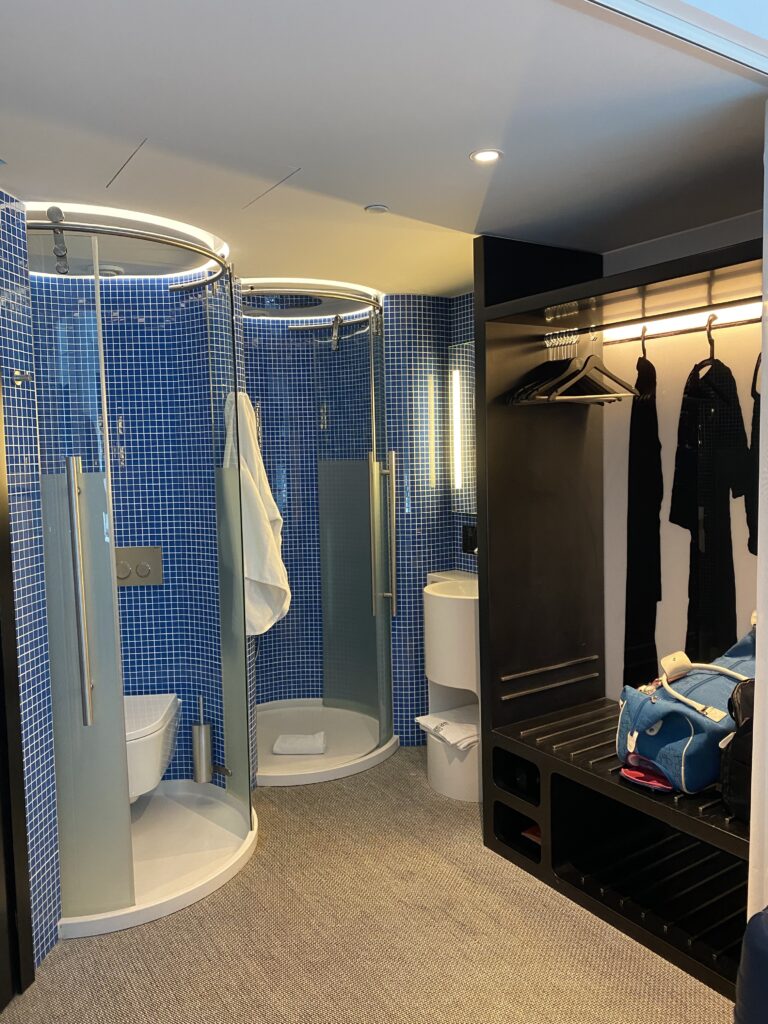 Inside of hotel room with two glass pods for a shower and toilet, a sink area, and clothing dresser.