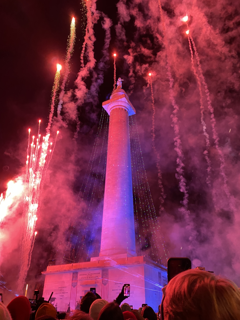 Washington monument in Baltimore Maryland draped in holiday lights and fireworks going up around it