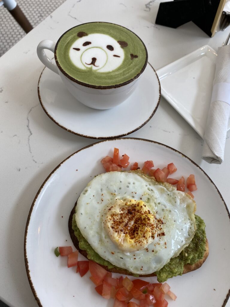Latte mug with latte art of a panda and a plate of avocado toast with a fried egg on a cafe table.