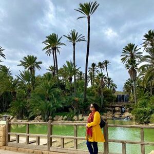 Woman in yellow jacket in an Alicante, Spain palm tree park and a lake behind her