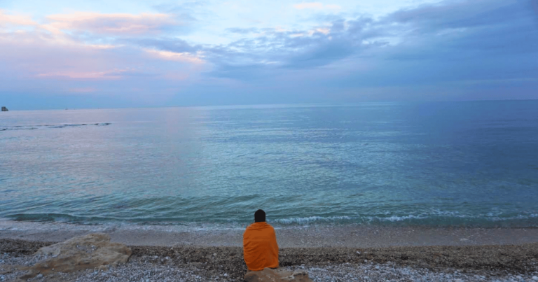 Woman wrapped in orange blanket sitting on a white pebble beach looking at a blue, pink, and purple sunset over the Mediterranean Sea