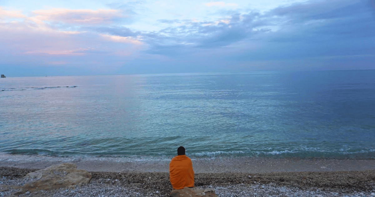 Woman wrapped in orange blanket sitting on a white pebble beach looking at a blue, pink, and purple sunset over the Mediterranean Sea
