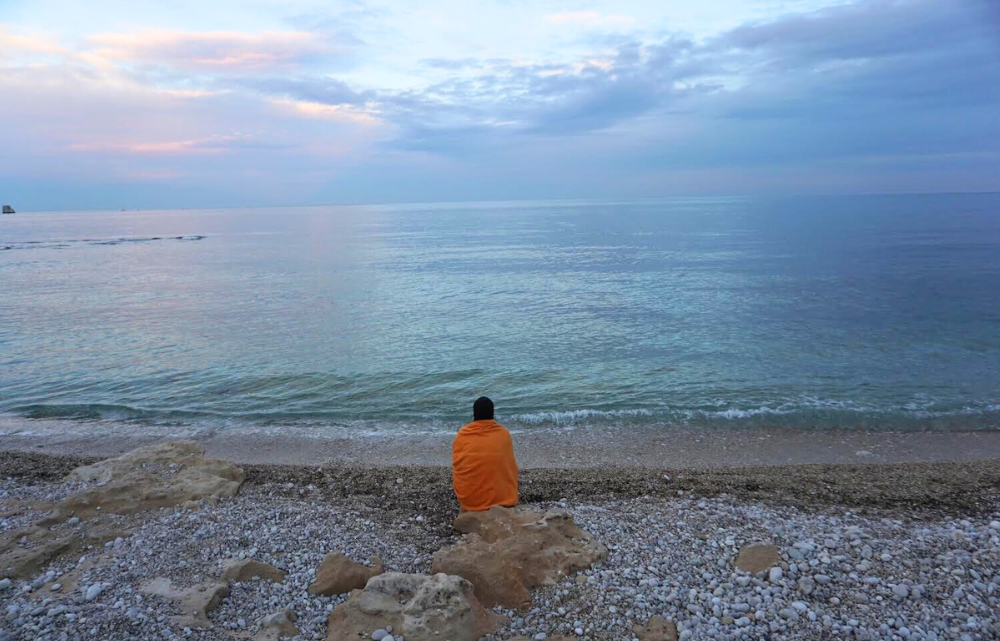 Woman wrapped in orange blanket sitting on a white pebble beach looking at a blue, pink, and purple sunset over the Mediterranean Sea