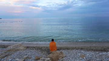Woman wrapped in orange blanket sitting on a white pebble beach looking at a blue, pink, and purple sunset over the Mediterranean Sea