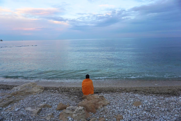 Woman wrapped in orange blanket sitting on a white pebble beach looking at a blue, pink, and purple sunset over the Mediterranean Sea