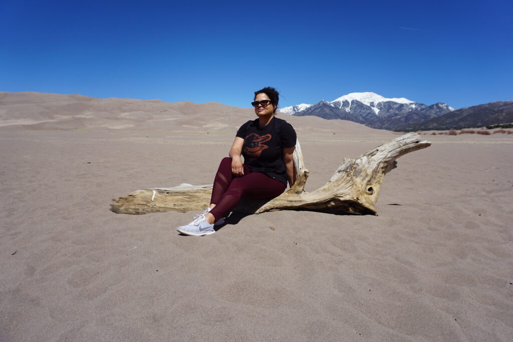 Woman sitting on dry log with snow-capped mountains and sand dunes in background