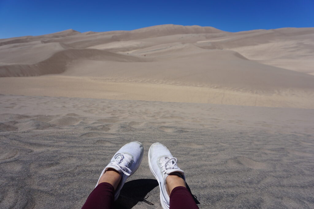 Feet in white sneakers against a backdrop of Colorado sand dunes