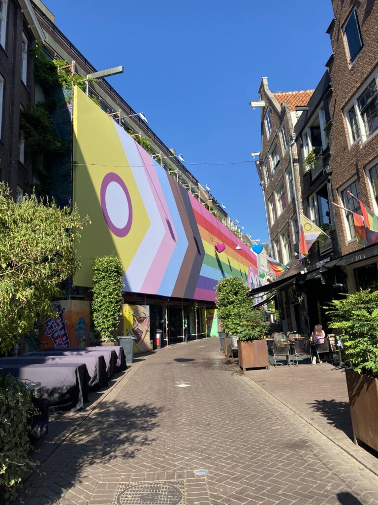 Amsterdam city street with a large LGBTQIA+ flag on the side of a row of buildings