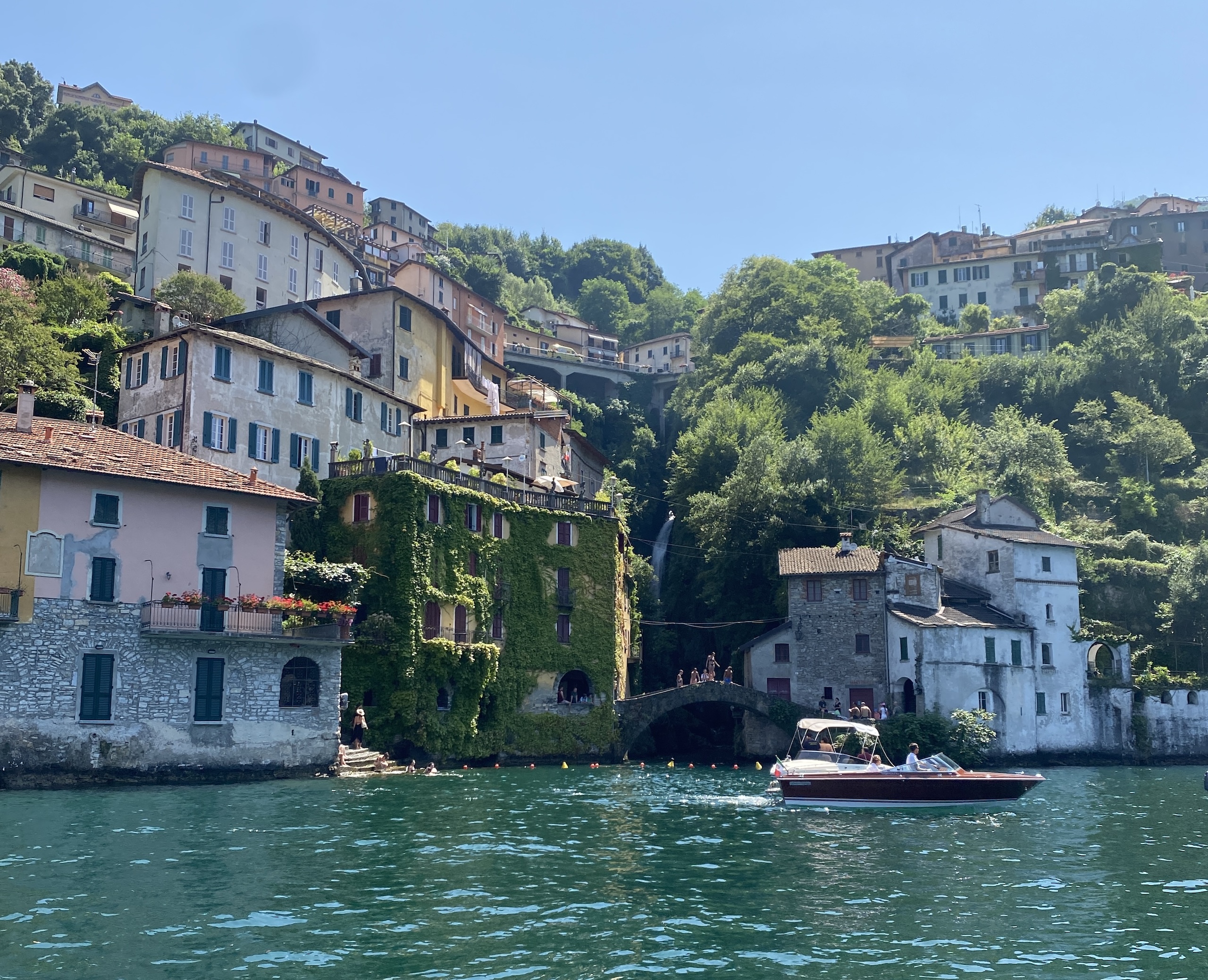 View from the water of Nesso, a Lake Como Italy town with a waterfall and bridge where people are jumping off into the water.