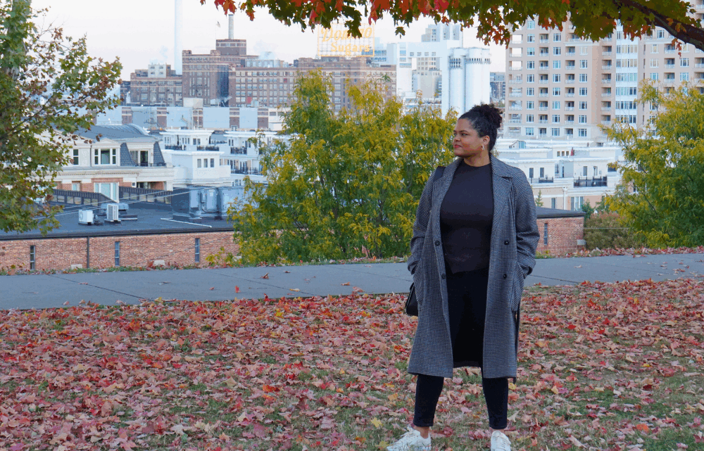 Woman in park overlooking a city with red and orange autumn leaves on the ground and trees with fall leaf colors