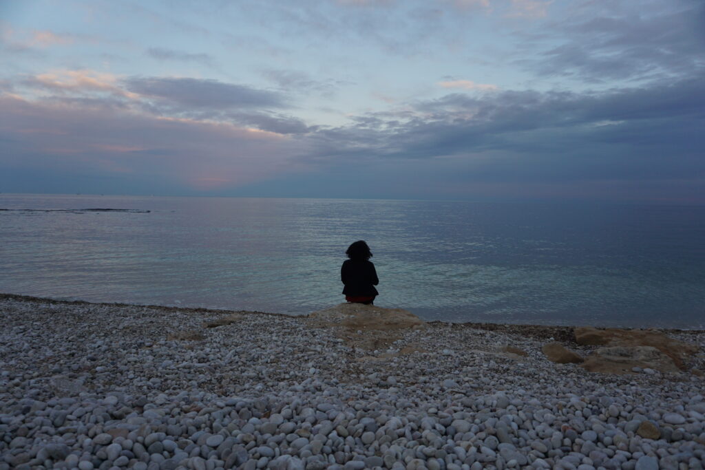 Woman sitting on a rock on a white pebble beach with a pink and light blue sunset in the sky