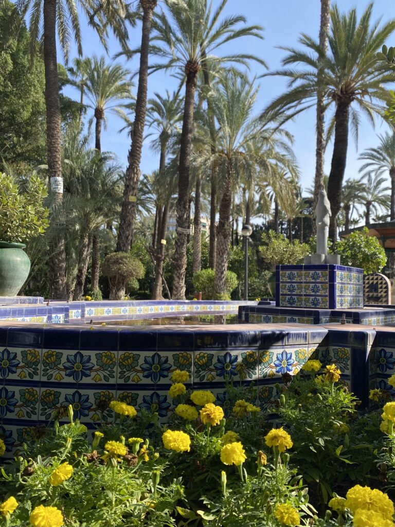 Mediterranean-tiled fountain with yellow flowers in the foreground and tall palm trees in the background