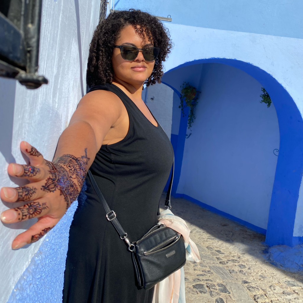 Woman in black dress showing her hand and arm decorated with henna against a blue wall outdoors
