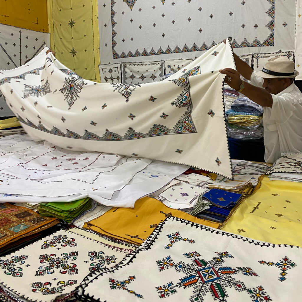Two men holding up a large traditionally embroidered tablecloth in a textile store in Morocco