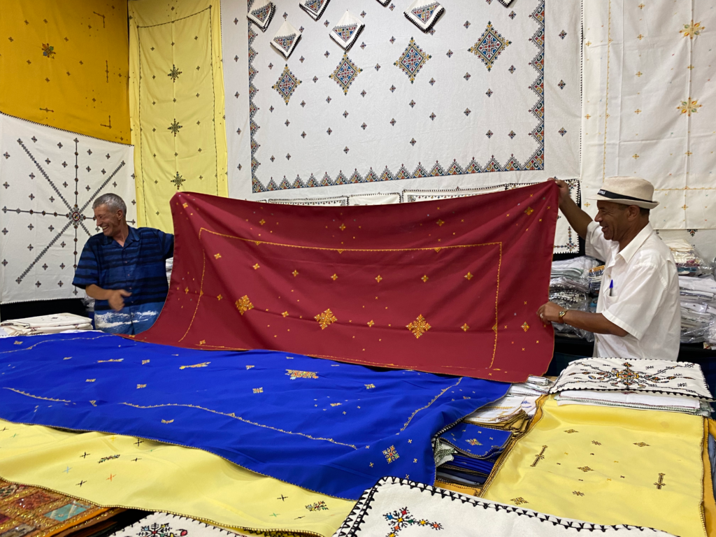 Two men holding up a large red traditionally embroidered tablecloth in a textile store in Morocco