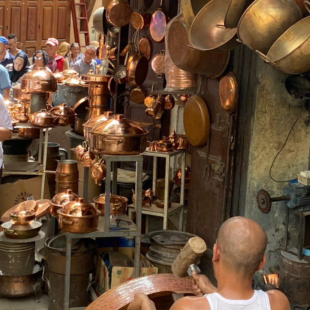Copper metalware hanging on display as a seated man hammers a piece of metal