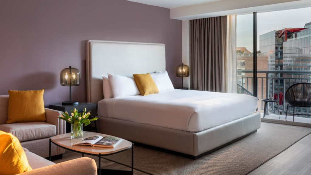 Inside of hotel room with one queen bed, two armchairs, and floor to ceiling windows looking out on city buildings