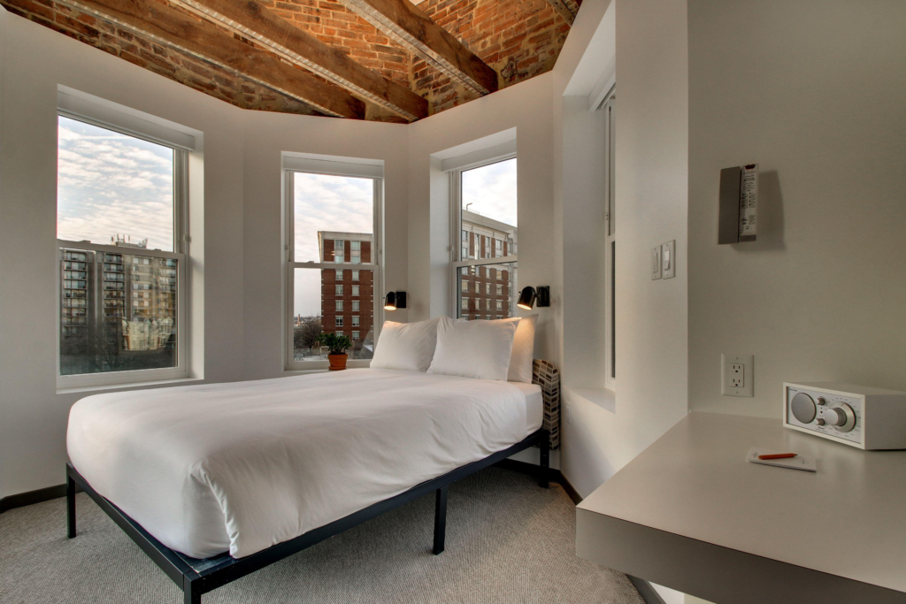 Inside of a small hotel room with a simple bed, small desk, and four windows with a Washington DC city view.