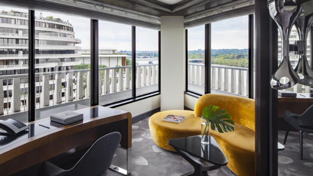 Watergate Hotel guest room with a desk and chair and yellow curved sofa next to large windows looking out on the Potomac River and other wing of the hotel.