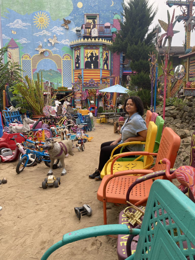 Woman sitting on an outdoors chair in a row of multicolored chairs with quirky decorations across the sand-covered ground